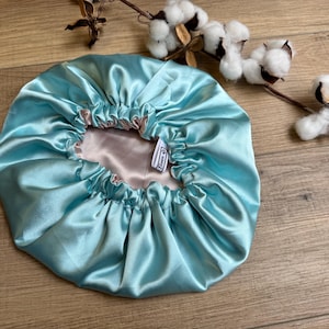 Könnte beinhalten: Eine hellblaue Satin-Haarmütze mit rosa Satin-Futter. Die Mütze ist oben gerafft und hat ein kleines weißes Etikett.