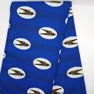 May include: Blue fabric featuring a repeating pattern of black and gold birds within white ovals. The background has thin, horizontal blue stripes. The fabric is folded, showing multiple layers. This textile is suitable for sewing projects.