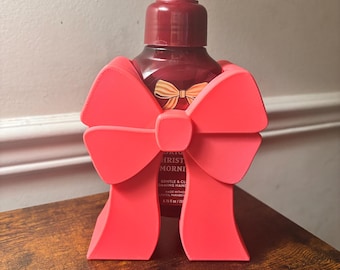 Puede incluir: Un dispensador de jabón de manos rojo con un soporte en forma de lazo rojo a juego. El dispensador tiene una bomba y una etiqueta con texto. El soporte de lazo es un accesorio decorativo. El dispensador de jabón está sobre una superficie de madera.
