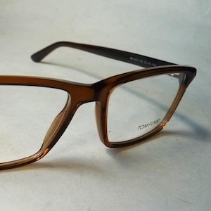 Może przedstawiać: Brązowe prostokątne okulary z złotym akcentem na zauszniku. Okulary mają przezroczyste soczewki, a na soczewce widoczna jest nazwa marki "TOM FORD". Oprawki są w ciepłym brązowym kolorze.