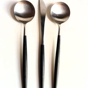 May include: A set of three stainless steel utensils: two soup spoons and a butter knife. Each piece has a black handle. The spoons have round bowls, and the knife has a rounded blade. The utensils are arranged on a white background.