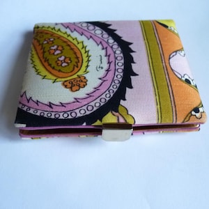 May include: A colorful wallet with a paisley pattern in shades of pink, orange, yellow, and black. The wallet has a silver clasp and is closed. The design is a vintage style.