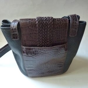 May include: A brown leather handbag with a black leather flap and a braided strap. The bag has a crocodile-embossed design.