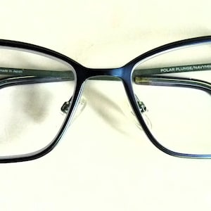 May include: Black cat-eye glasses with clear lenses and a dark green frame. The glasses have the text "POLAR PLUNGE/NAVY/HT" and "54□18 145" printed on the right arm.