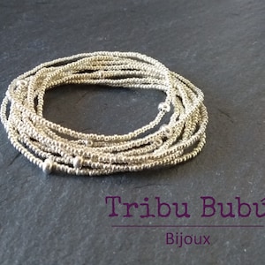 May include: A silver beaded bracelet with small silver beads. The bracelet is made of multiple strands of beads and is coiled up on a dark gray surface. The text "Tribu Bubú Bijoux" is visible below the bracelet.