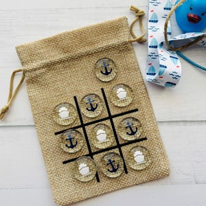 May include: A burlap drawstring bag with a tic-tac-toe game featuring clear glass game pieces with anchor and sailboat designs. A blue rubber duck, nautical ribbon, and twine are also present.