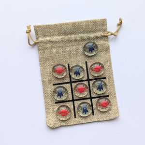 May include: A burlap drawstring bag with a tic-tac-toe board printed on the front. The board is filled with glass game pieces with red planets and blue spaceships.