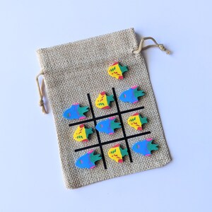 May include: A burlap drawstring bag with a tic-tac-toe game printed on it. The game pieces are colorful fish with yellow, blue, and red accents.
