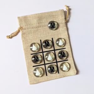 May include: A burlap bag with a tic-tac-toe board printed on it. The board is filled with glass game pieces with images of a wedding dress and a tuxedo.