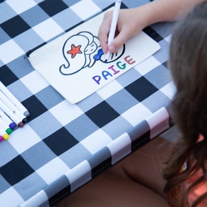 Puede incluir: Una bolsa de lona blanca con un dise&ntilde;o de sirena y el nombre "PAIGE" escrito en letras de colores. Una persona est&aacute; coloreando la bolsa con marcadores sobre una mesa a cuadros blancos y negros.