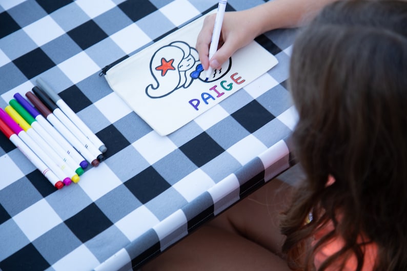 Puede incluir: Una bolsa de lona blanca con un dise&ntilde;o de sirena y el nombre "PAIGE" escrito en letras de colores. Una persona est&aacute; coloreando la bolsa con marcadores sobre una mesa a cuadros blancos y negros.