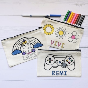 May include: Three white canvas zippered pouches with different designs for coloring. The first pouch has a unicorn with a rainbow and clouds, the second pouch has a sun, a daisy, and a smaller flower, and the third pouch has a video game controller. Each pouch has a name written on it: Lexi, Vivi, and Remi.