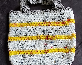May include: A white crocheted tote bag with yellow stripes. The bag is made from recycled plastic and has a black and white crocheted handle.