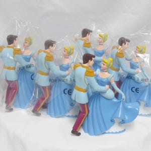 May include: A collection of Cinderella figurines in plastic packaging. Each figurine depicts Cinderella in a blue gown dancing with Prince Charming. The prince wears a light blue jacket with gold accents and burgundy pants. The figurines are marked "FOR AGES 3 AND UP".