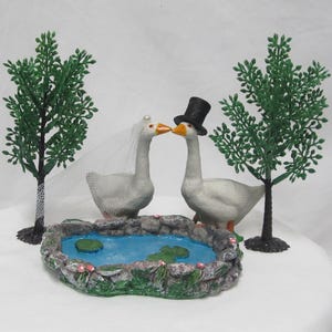 May include: A wedding cake topper featuring two white geese kissing, one wearing a top hat and the other a veil. A small pond with lily pads and two green trees flank the geese. The pond is surrounded by a stone border.