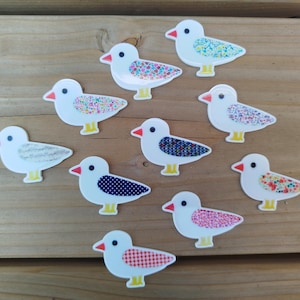 Op de afbeelding: Een verzameling van negen platte vogelvormige knopen, elk met een wit lichaam, een kleine oranje snavel, een zwart oog en gele pootjes. Elke vogel heeft een uniek vleugelpatroon, waaronder bloemmotieven, stippen, ruitjes en geometrische ontwerpen.