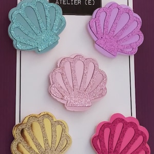 May include: Set of five glitter seashell hair clips in various colours, including teal, purple, pink, gold, and hot pink. The clips are displayed on a white card with the text "ATELIER (E)" and "les & Cie/aime.annuel."
