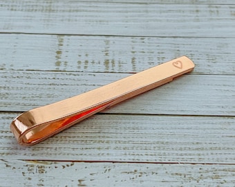 May include: A rose gold tie bar with a heart engraved on the top.
