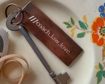以下が含まれることがあります： 「//coach.law.love」の文字が入った銅色の長方形キーホルダーが、アンティークキーと一緒に花柄のプレートに置かれています。 プレートはクリーム色の背景に、黄色、オレンジ、青の花のデザインが施されています。