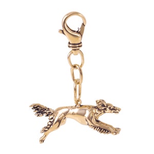 May include: A gold-colored metal charm featuring a running dog design. The charm is attached to a lobster clasp and chain. The dog is detailed with textured fur and a flowing tail. The charm is likely for a keychain or jewelry.
