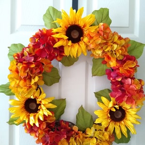 Puede incluir: Una vibrante corona circular con girasoles y hortensias en tonos amarillos, naranjas y rojos, acentuada por hojas verdes. La corona se exhibe contra una puerta blanca, creando una estética otoñal alegre.