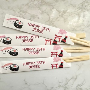 Puede incluir: Cuatro envoltorios de palillos de papel blanco con un dise&ntilde;o de sushi rojo y negro. Los envoltorios tienen el texto "Happy 35th Jesse" impreso en ellos. Los envoltorios est&aacute;n doblados alrededor de palillos de madera.
