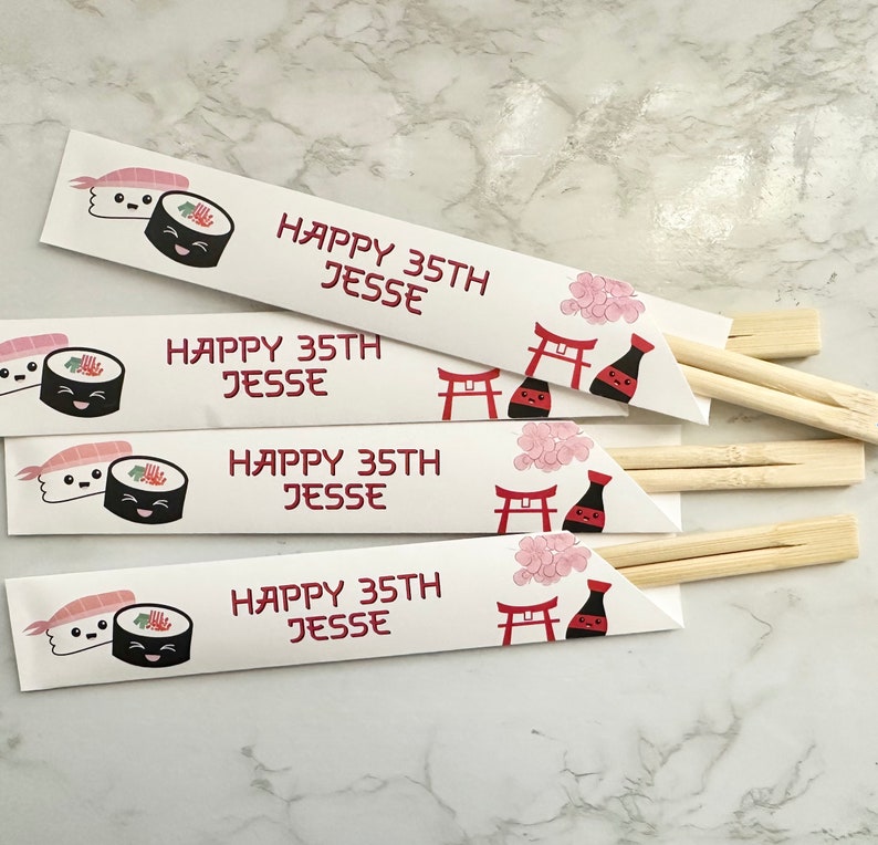 Puede incluir: Cuatro envoltorios de palillos de papel blanco con un dise&ntilde;o de sushi rojo y negro. Los envoltorios tienen el texto "Happy 35th Jesse" impreso en ellos. Los envoltorios est&aacute;n doblados alrededor de palillos de madera.