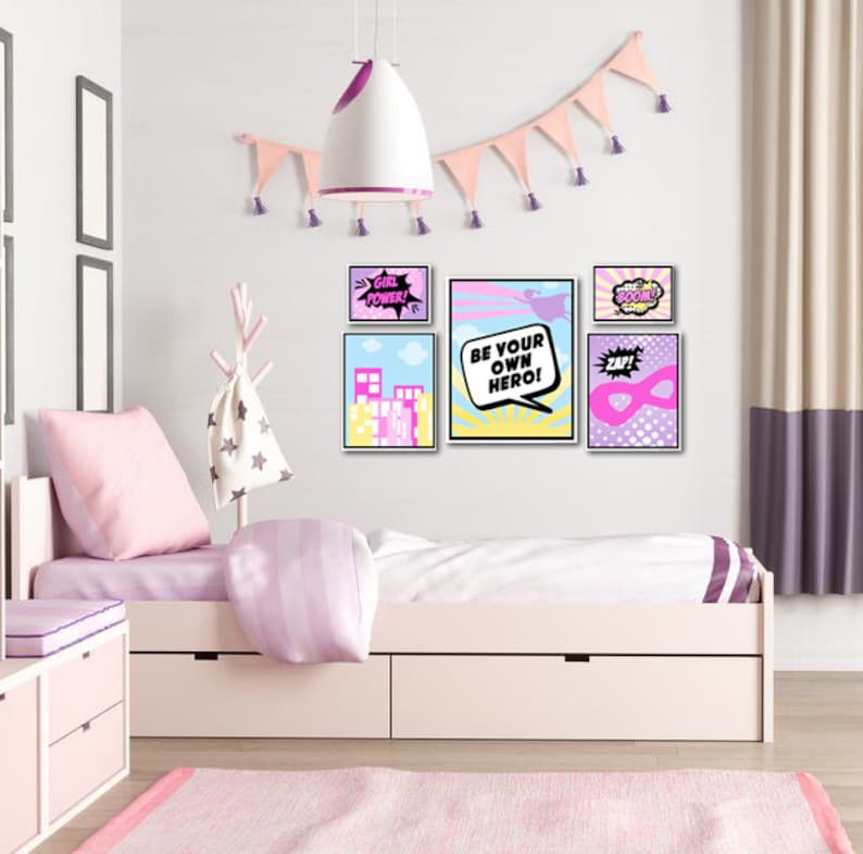 May include: A set of six colorful prints with a superhero theme. The prints feature phrases like "Girl Power!" and "Be Your Own Hero!" and are displayed on a white wall above a pink bed.