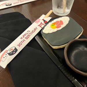 Puede incluir: Un favor de fiesta de cumplea&ntilde;os con tem&aacute;tica de sushi en blanco y negro con el texto "Karl's 30th Birthday" en un porta palillos blanco. El favor incluye una galleta de sushi en blanco y negro con un camar&oacute;n rojo y un huevo amarillo en la parte superior.