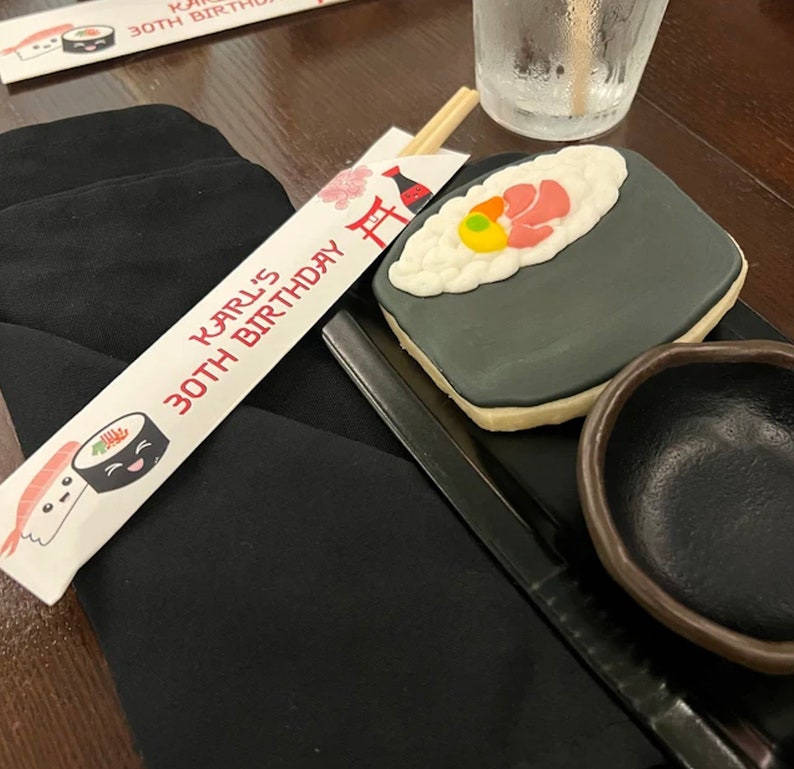 Puede incluir: Un favor de fiesta de cumplea&ntilde;os con tem&aacute;tica de sushi en blanco y negro con el texto "Karl's 30th Birthday" en un porta palillos blanco. El favor incluye una galleta de sushi en blanco y negro con un camar&oacute;n rojo y un huevo amarillo en la parte superior.