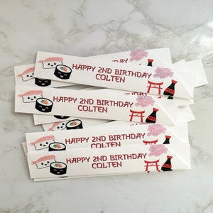 Puede incluir: Una pila de tarjetas rectangulares blancas con un dise&ntilde;o de sushi y el texto "Happy 2nd Birthday Colten" impreso en rojo. Las tarjetas presentan un rollo de sushi de dibujos animados, una gamba de dibujos animados, una botella de salsa de soja de dibujos animados y un templo japon&eacute;s de dibujos animados.