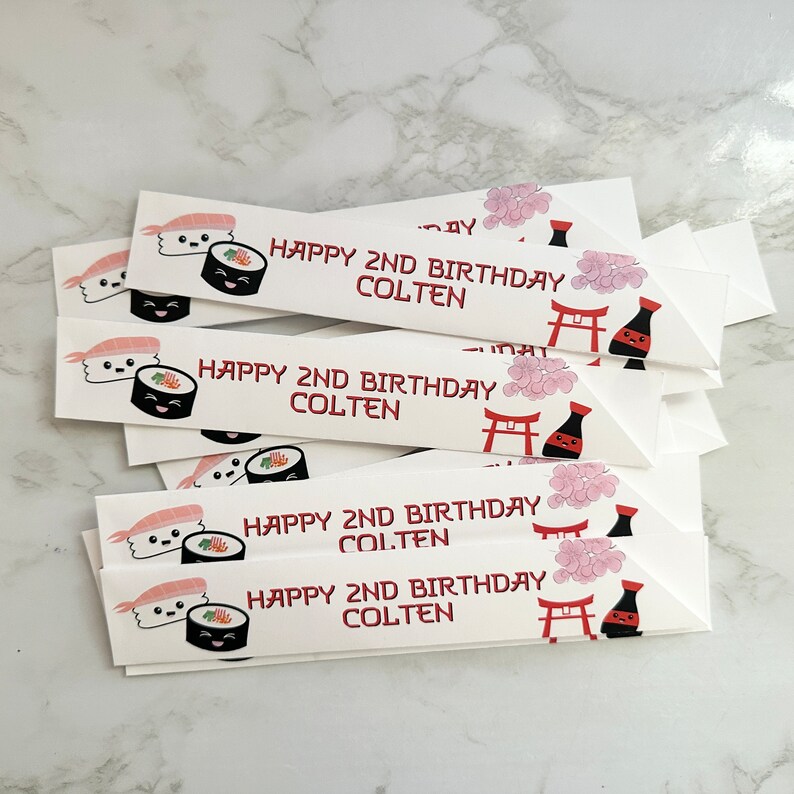 Puede incluir: Una pila de tarjetas rectangulares blancas con un dise&ntilde;o de sushi y el texto "Happy 2nd Birthday Colten" impreso en rojo. Las tarjetas presentan un rollo de sushi de dibujos animados, una gamba de dibujos animados, una botella de salsa de soja de dibujos animados y un templo japon&eacute;s de dibujos animados.