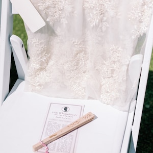 May include: A white chair supports a cream-coloured, lacy garment with floral embroidery. A blank tag hangs from the garment. A wooden fan and a printed paper with the text "Lady Whistledown's Society Papers" rest on the chair.