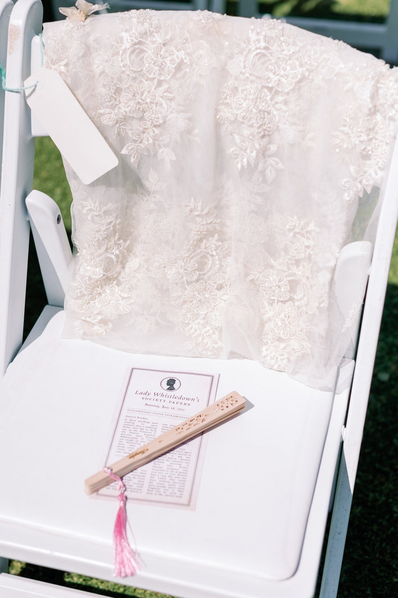 May include: A white chair supports a cream-coloured, lacy garment with floral embroidery. A blank tag hangs from the garment. A wooden fan and a printed paper with the text "Lady Whistledown's Society Papers" rest on the chair.