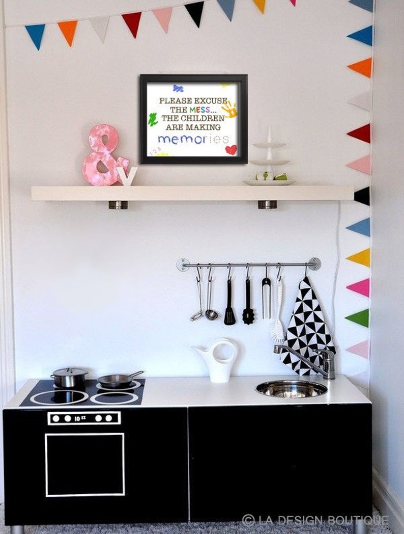 May include: A black framed sign with the text "PLEASE EXCUSE THE MESS... THE CHILDREN ARE MAKING memories" in colorful letters. The sign is hanging on a white shelf above a black and white play kitchen with a sink, stove, and oven.