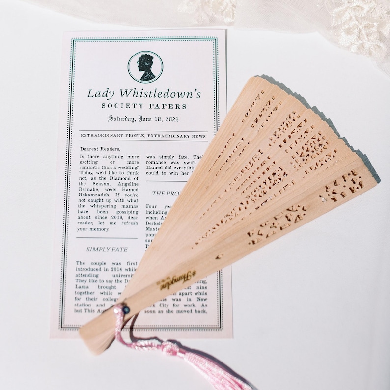May include: A light-coloured wooden hand fan with intricate cut-out designs, featuring a pink tassel. The fan is positioned in front of a printed paper with the text "Lady Whistledown's Society Papers".