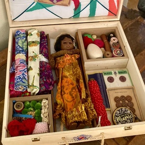 May include: A wooden box with a hinged lid and a rope handle. The box is filled with various sewing supplies, including fabric scraps, buttons, needles, thread, and a doll. The lid of the box is lined with a fabric featuring a floral pattern and a green grid.