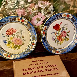 Puede incluir: Dos platos de porcelana vintage con diseños florales y bordes azules. Los platos se muestran con una caja que dice "UNBREAKABLE PORCELAIN COLOR MATCHING PLATES IMPORTED FROM HOLLAND". La caja recomienda los platos para decoraciones de pared, cuencos de dulces y platos de aperitivos de TV.