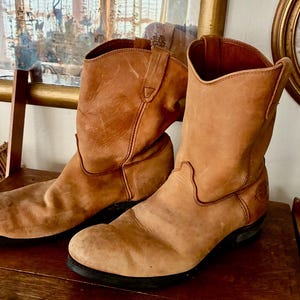 May include: A pair of brown leather cowboy boots with a worn, distressed appearance. The boots have a mid-calf height, rounded toes, and a slight heel. They are placed on a wooden surface, showcasing their vintage style.