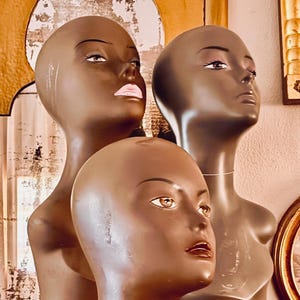 Könnte beinhalten: Drei Schaufensterpuppenköpfe mit braunen Hauttönen werden gezeigt. Die Köpfe haben bemalte Gesichtszüge, einschließlich Augen, Augenbrauen und Lippen. Die Schaufensterpuppen sind vor einem Spiegel und anderen dekorativen Gegenständen positioniert.