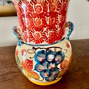 May include: A hand-painted ceramic vase with a wide opening and two small handles. The top section features a red floral pattern, while the lower section displays a cluster of blue grapes and colorful leaves. The rim is blue.