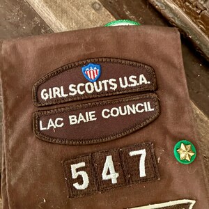 K&ouml;nnte beinhalten: Braune Girl Scout Weste mit gestickten Aufn&auml;hern. Aufn&auml;her sind unter anderem "Girl Scouts U.S.A.", "Lac Baie Council" und die Nummer "547". Zus&auml;tzliche Aufn&auml;her und eine gr&uuml;ne und goldene Anstecknadel sind ebenfalls sichtbar.