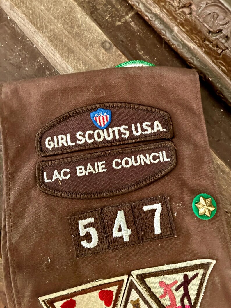 K&ouml;nnte beinhalten: Braune Girl Scout Weste mit gestickten Aufn&auml;hern. Aufn&auml;her sind unter anderem "Girl Scouts U.S.A.", "Lac Baie Council" und die Nummer "547". Zus&auml;tzliche Aufn&auml;her und eine gr&uuml;ne und goldene Anstecknadel sind ebenfalls sichtbar.