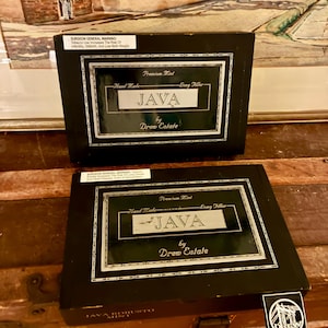 Op de afbeelding: Twee zwarte dozen met het woord "JAVA" in een decoratief kader. De dozen hebben de tekst "Premium Mint", "Hand Made" en "Long Filler". De dozen staan op een houten oppervlak.