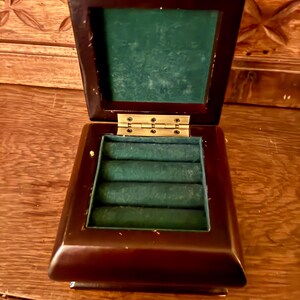 May include: Open, dark brown wooden jewelry box with a green velvet interior. The box has a hinged lid and brass-colored hinges. The interior features slots for rings. The box is sitting on a wooden surface.