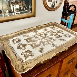May include: A rectangular serving tray with a scalloped edge and a metallic gold finish. The center features a white surface decorated with intricate, symmetrical brown scrollwork and floral patterns that extend across the entire base.