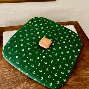 May include: A square, dark green lid featuring a repeating white floral pattern and a centered, light-colored wooden knob. The lid has rounded corners and a smooth finish, designed to fit securely onto a container or box for storage purposes.