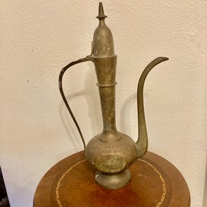 Puede incluir: Una tetera de latón vintage con intrincados diseños grabados. La tetera tiene un pico alto y delgado y un cuerpo redondeado. Se encuentra sobre una mesa de madera con un borde dorado.