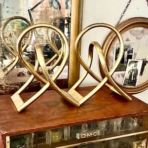 Puede incluir: Dos esculturas doradas abstractas en forma de cinta. Cada escultura tiene una base rectangular y un diseño en bucle. Se exhiben sobre una superficie de madera, con un espejo vintage y fotografías enmarcadas en el fondo.