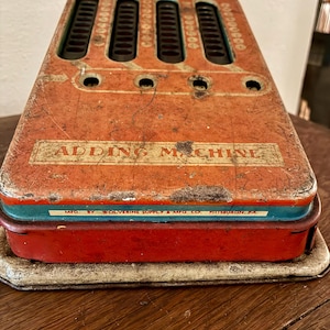 Op de afbeelding: Vintage rode en teal optelmachine met de tekst "ADDING MACHINE" en "MFD. BY WOLVERINE SUPPLY & MFG. CO. PITTSBURGH, PA." De machine heeft vier rijen nummer toetsen en is gemaakt van metaal.